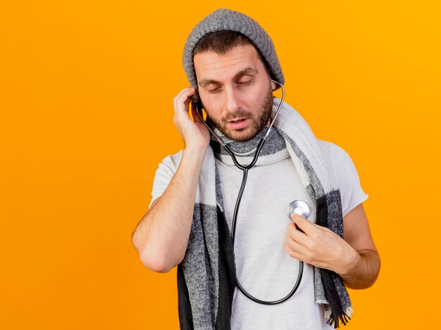 Con los ojos cerrados joven enfermo con gorro de invierno y bufanda escuchando los latidos de su propio corazón con estetoscopio aislado sobre fondo amarillo