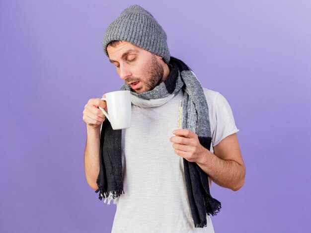 Con los ojos cerrados joven enfermo con sombrero de invierno con bufanda sosteniendo una taza de té y un termómetro aislado sobre fondo púrpura