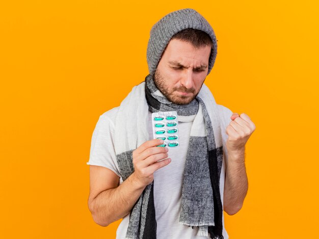 Con los ojos cerrados joven enfermo vestido con gorro de invierno y bufanda sosteniendo pastillas y mostrando sí gesto aislado sobre fondo amarillo