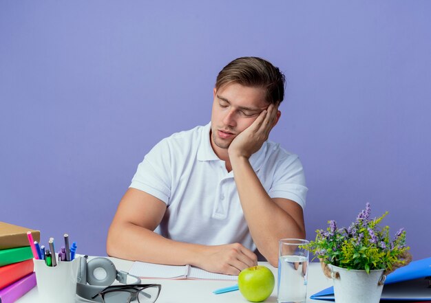 Con los ojos cerrados, joven y guapo estudiante sentado en un escritorio con herramientas escolares mostrando gesto de sueño aislado en azul