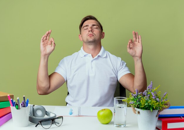 Con los ojos cerrados, joven y guapo estudiante sentado en un escritorio con herramientas escolares que muestran gesto de meditación aislado en verde oliva