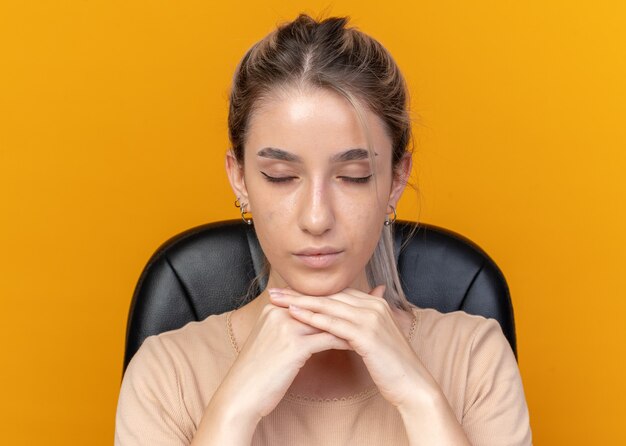 Con los ojos cerrados joven hermosa se sienta a la mesa con herramientas de maquillaje poniendo la mano debajo de la barbilla aislada sobre fondo naranja