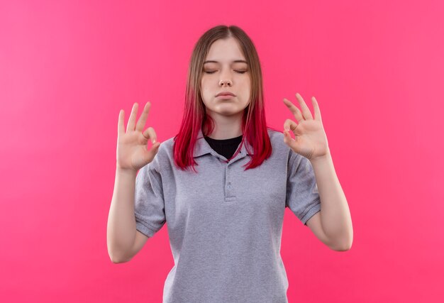 Con los ojos cerrados joven hermosa vestida con camiseta gris que muestra el gesto okey en la pared rosa aislada