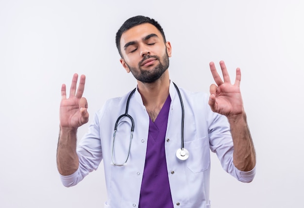 Con los ojos cerrados joven médico con estetoscopio bata médica mostrando gesto okey en blanco aislado