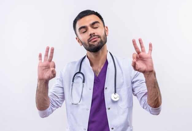 Con los ojos cerrados joven médico con estetoscopio bata médica mostrando gesto okey en blanco aislado