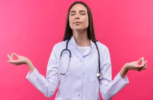 Con los ojos cerrados joven médico con estetoscopio bata médica mostrando gesto okey en pared rosa aislada