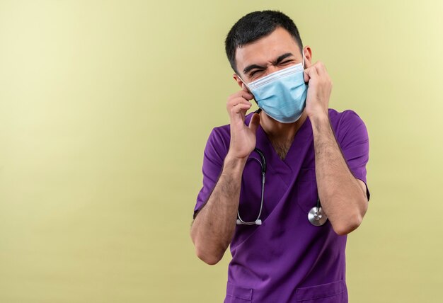 Con los ojos cerrados joven médico vistiendo ropa de cirujano púrpura y estetoscopio máscara médica orejas cerradas en pared verde aislada