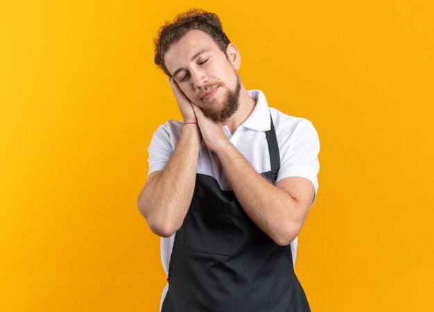 Con los ojos cerrados joven peluquero vistiendo uniforme mostrando gesto de sueño aislado sobre fondo amarillo