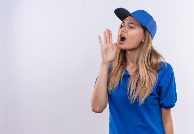 Con los ojos cerrados joven repartidora vestida con uniforme azul y gorra llamando a alguien aislado en la pared blanca con espacio de copia