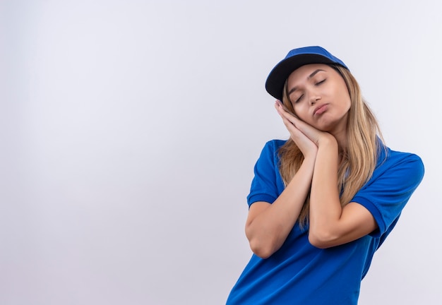 Con los ojos cerrados joven repartidora vestida con uniforme azul y gorra mostrando gesto de sueño aislado en blanco