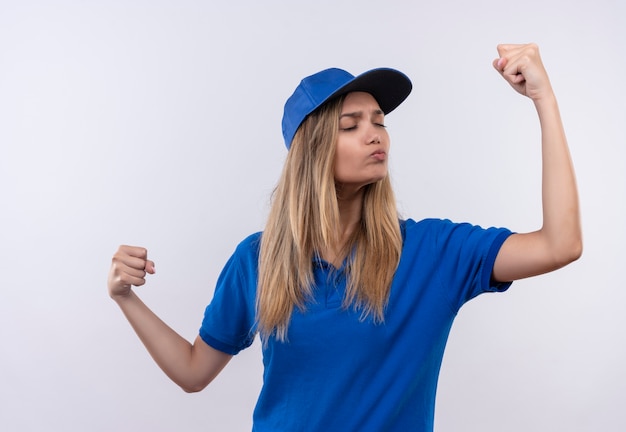 Foto gratuita con los ojos cerrados joven repartidora vestida con uniforme azul y gorra que muestra un gesto fuerte aislado en la pared blanca con espacio de copia