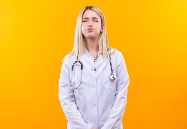 Con los ojos cerrados, médico joven con estetoscopio en bata médica mostrando gesto de beso en la pared amarilla aislada