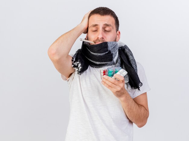 Foto gratuita con los ojos cerrados molesto joven enfermo vistiendo bufanda envuelta en plaid sosteniendo pastillas
