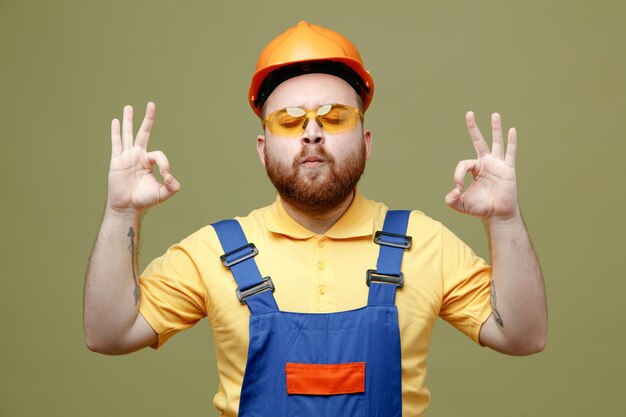 Con los ojos cerrados mostrando meditación joven constructor hombre en uniforme aislado sobre fondo verde