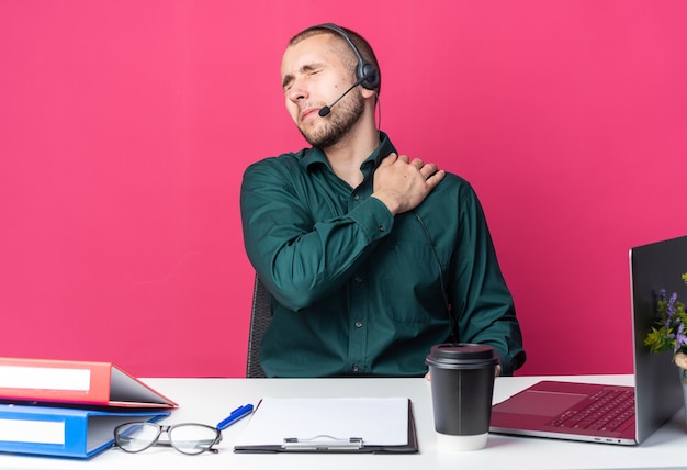 Con los ojos cerrados, el operador del centro de llamadas masculino joven con auriculares sentado en un escritorio con herramientas de oficina agarró el hombro dolorido