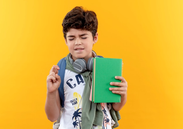Con los ojos cerrados, pequeño niño de escuela preocupado con mochila y auriculares sosteniendo el libro y cruzando los dedos