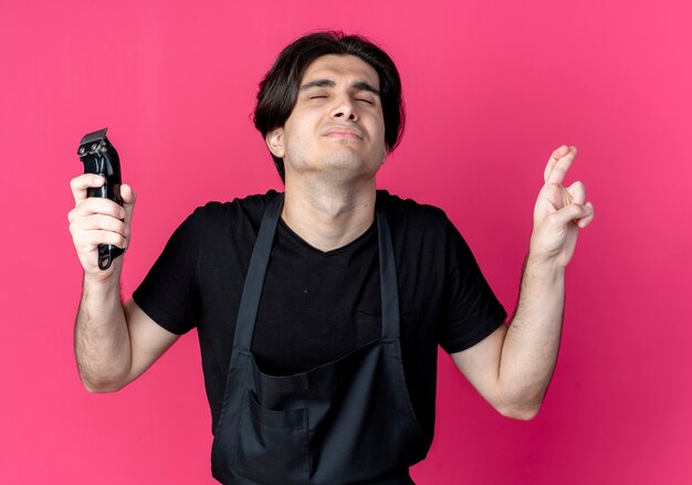 Con los ojos cerrados preocupado joven guapo barbero en uniforme sosteniendo cortapelos y cruzando los dedos aislados en rosa