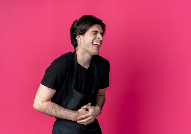 Con los ojos cerrados riendo joven barbero guapo en uniforme agarró el estómago aislado en rosa