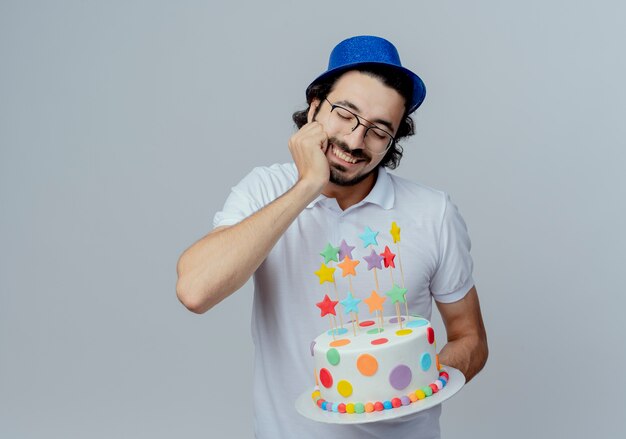 Con los ojos cerrados sonriendo guapo con gafas y sombrero azul sosteniendo la torta y poniendo la mano debajo de la barbilla aislado en blanco