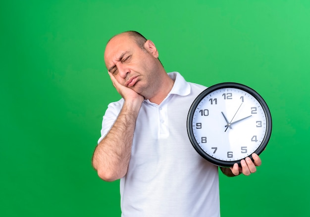 Con los ojos cerrados, triste, casual, hombre maduro, tenencia, reloj de pared, y, poniendo, mano, en, mejilla, aislado, en, pared verde