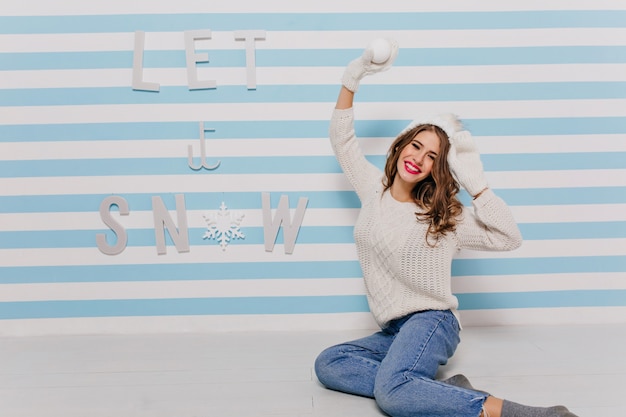 Los ojos de la linda chica brillan de felicidad en previsión del divertido juego de bolas de nieve. Foto de modelo en jeans en pared rayada