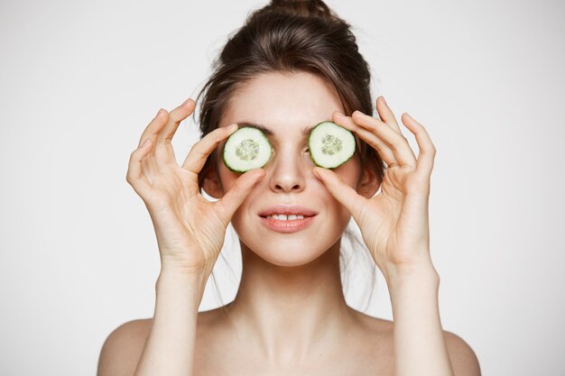 Ojos de ocultación sonrientes hermosos jovenes desnudos de la muchacha detrás de rebanadas del pepino sobre el fondo blanco. Belleza, cuidado de la piel y cosmetología.