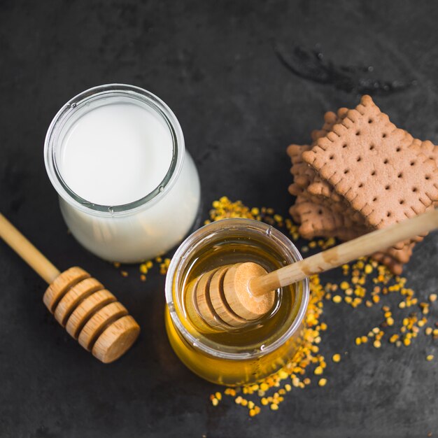 Olla de leche; tarro de miel; polen de abeja y pila de galletas en el telón de fondo con textura