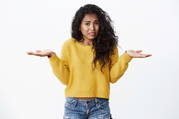 Oops girl dice lo siento Linda mujer afroamericana tonta cabello negro rizado encogiéndose de hombros manos extendidas de lado frunciendo el ceño y llorando como si se sintiera preocupada disculpándose por cometer un error stand fondo blanco