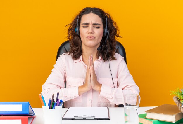 Operador de centro de llamadas femenino bastante caucásico ansioso en auriculares sentado en el escritorio con herramientas de oficina y rezando aislado en la pared naranja