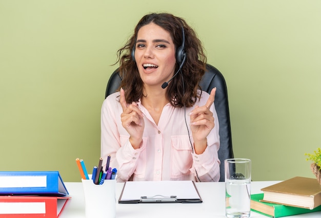 Operador de centro de llamadas femenino bastante caucásico complacido en auriculares sentado en el escritorio con herramientas de oficina apuntando hacia arriba aislado en la pared verde