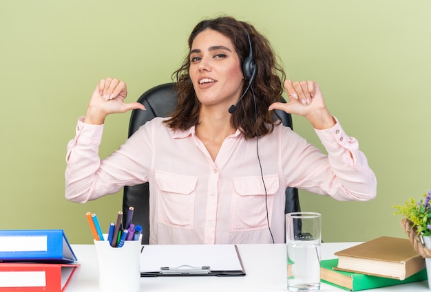 Foto gratuita operador de centro de llamadas femenino bastante caucásico complacido en auriculares sentado en el escritorio con herramientas de oficina apuntando a sí misma aislada en la pared verde