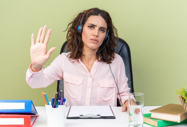 Operador de centro de llamadas femenino bastante caucásico disgustado en auriculares sentado en el escritorio con herramientas de oficina gesticulando señal de parada de mano