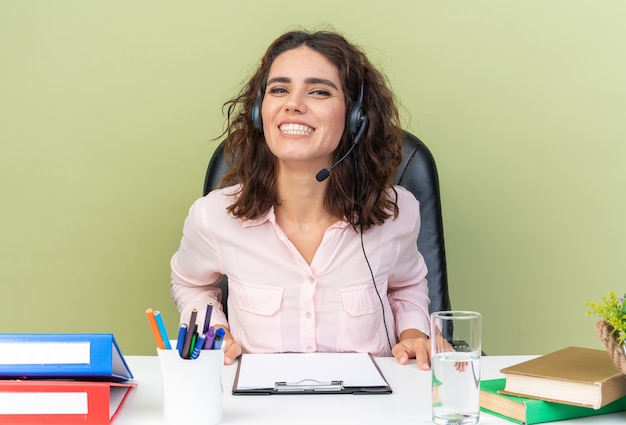 Foto gratuita operador de centro de llamadas femenino bastante caucásico sonriente en auriculares sentado en el escritorio con herramientas de oficina aisladas en la pared verde