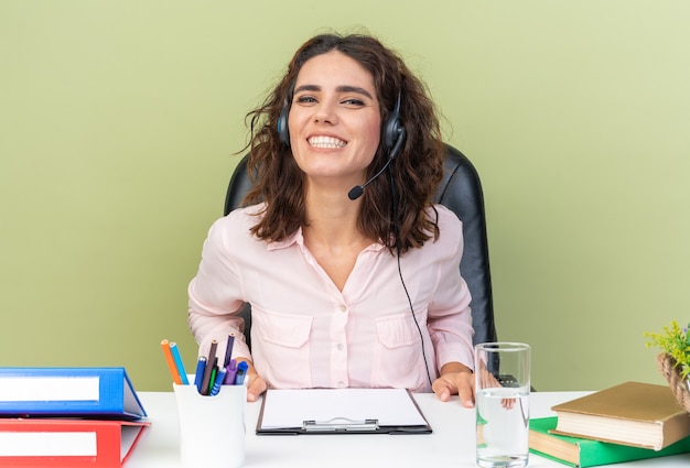 Operador de centro de llamadas femenino bastante caucásico sonriente en auriculares sentado en el escritorio con herramientas de oficina aisladas en la pared verde