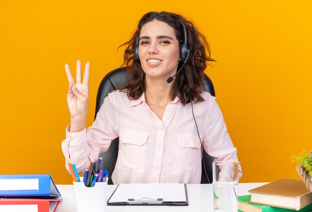 Operador de centro de llamadas femenino bastante caucásico sonriente en auriculares sentado en el escritorio con herramientas de oficina gesticulando tres con los dedos