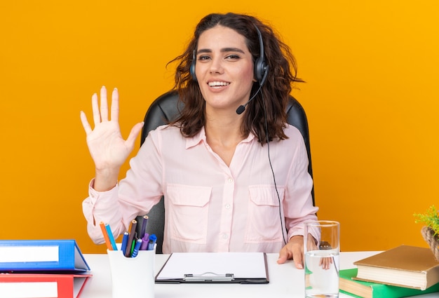 Foto gratuita operador de centro de llamadas femenino bastante caucásico sonriente en auriculares sentado en el escritorio con herramientas de oficina manteniendo la mano abierta aislada en la pared naranja