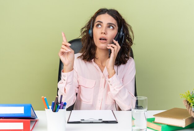 Operador de centro de llamadas femenino bastante caucásico sorprendido en auriculares sentado en el escritorio con herramientas de oficina hablando por teléfono mirando y apuntando hacia arriba aislado en la pared verde