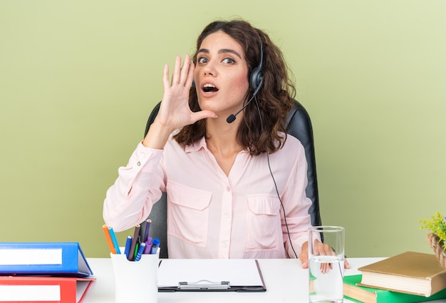 Operador de centro de llamadas femenino bastante caucásico sorprendido en auriculares sentado en el escritorio con herramientas de oficina manteniendo la mano cerca de la boca