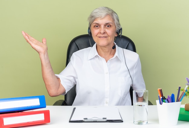 Operador de centro de llamadas femenino caucásico complacido en auriculares sentado en el escritorio con herramientas de oficina manteniendo la mano abierta