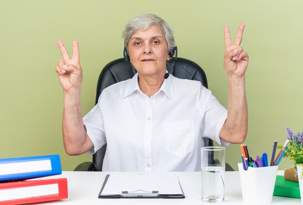 Operador de centro de llamadas femenino caucásico confiado en auriculares sentado en el escritorio con herramientas de oficina gesticulando signo de victoria aislado en la pared verde