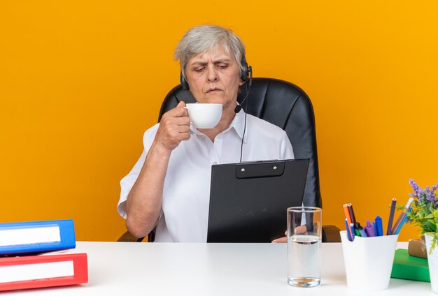 Operador de centro de llamadas femenino caucásico confiado en auriculares sentado en el escritorio con herramientas de oficina sosteniendo la taza y mirando el portapapeles