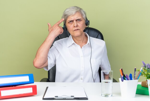 Operador de centro de llamadas femenino caucásico disgustado en auriculares sentado en el escritorio con herramientas de oficina apuntando a su cabello