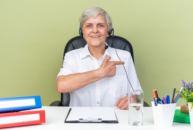 Operador de centro de llamadas femenino caucásico sonriente en auriculares sentado en el escritorio con herramientas de oficina apuntando al lado aislado en la pared verde