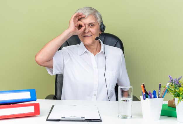 Operador de centro de llamadas femenino caucásico sonriente en auriculares sentado en el escritorio con herramientas de oficina a través de los dedos aislados en la pared verde