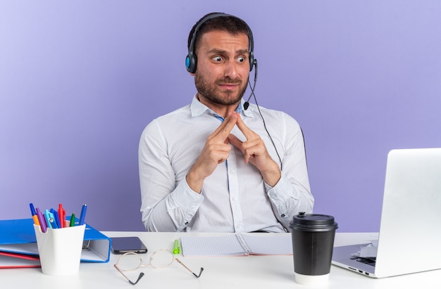 Foto gratuita operador de centro de llamadas masculino joven asustado con auriculares sentado a la mesa con herramientas de oficina mirando portátil