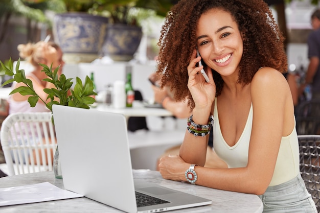 Operador de llamada mujer hermosa feliz de piel oscura para recibir consultany, utiliza dispositivos electrónicos, descansa en un acogedor café.