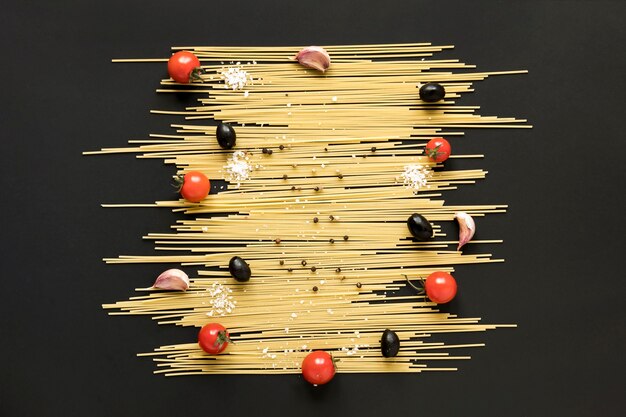 Opinión de alto ángulo de las pastas crudas de los espaguetis; tomate cherry; Aceituna negra y pimienta negra dispuestas en superficie negra.