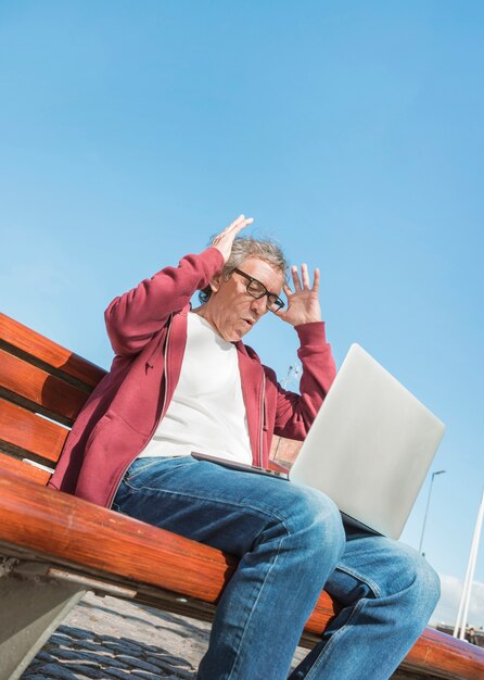 Opinión de ángulo bajo del hombre mayor con la computadora portátil en su regazo contra el cielo azul