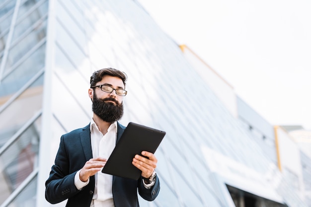 Opinión de ángulo bajo del hombre de negocios que mira la tableta digital que está parada fuera del edificio de oficinas