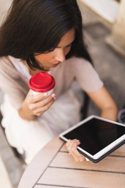 Opinión superior una muchacha que mira la tableta digital que sostiene la taza de café